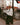 A person wearing a Rowing Blazers x Paddington™ Paddington Oxford Button Down White shirt, a dark necktie, and green plaid trousers stands by a London Underground station entrance.
