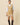 A person wearing a Women’s Cotton Blend Cable Dress Camel in a camel-colored V-neck cricket sweater style with striped trim over a white collared shirt, paired with tall brown boots.