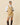 A model wears a Rowing Blazers Women’s Cotton Blend Cable Dress Camel, a tan sleeveless cricket sweater dress with striped trim over a white collared shirt, paired with dark tall boots.