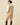 A person wearing a Women’s Cotton Blend Cable Dress Camel over a white collared shirt with striped trim at the neck and hem, paired with dark brown knee-high boots.