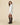 A person wears a white Rowing Blazers Women’s Button Down Pleated Shirt Dress in 100% cotton with a pleated skirt, black tights, and brown knee-high boots.