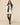A model wears a Women’s Button Down Pleated Shirt Dress White in 100% cotton with a dark brown cable-knit sweater draped over her shoulders and dark brown knee-high boots.