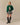 A model wears a dark green Rowing Blazers x Babar Babar Jardinier Cashmere Sweater featuring an intarsia gardening motif, paired with a black and white plaid pleated mini skirt and black knee-high boots.