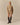 A woman stands against a beige background wearing Rowing Blazers Ollie Herringbone Casual Shorts Camel, a tan zip-up jacket, and tall dark brown boots.