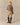 A person with long blonde hair, seen from behind, wears a patterned sweater, Rowing Blazers Ollie Herringbone Casual Shorts Camel, and knee-high dark brown boots against a beige background.