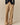 A person wears the Rowing Blazers Cath Herringbone Pant Camel with a drawstring waist, paired with a dark navy cardigan and black open-toe shoes, standing on a neutral background, hands in pockets.