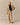 A woman stands against a beige background wearing the Cath Herringbone Pant Camel by Rowing Blazers, styled with a tan oversized jacket, black top, and open-toed heels. She faces forward with a neutral expression.