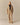 A person stands facing forward in a beige jacket and Rowing Blazers Cath Herringbone Pant Camel with a drawstring waist, dark cropped top, and open-toe sandals against a plain light background.