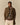 A man in a brown checked coat, pink shirt, beige pants, and the Rowing Blazers Donegal Fair Isle Crew Sweater Green stands with hands in pockets against a plain beige background.