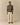 A person stands facing forward, wearing the Rowing Blazers Donegal Fair Isle Crew Sweater Green over a pink shirt, cream-colored pants, and brown shoes against a plain beige background.