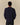 A person with short curly hair models the Cotton NY Hot Dog Sweater Navy by Rowing Blazers, paired with dark pants, standing against a plain beige background facing away from the camera.