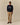 A man wears a navy blue Rowing Blazers x Paddington™ Paddington Cotton Text Sweater Navy embroidered with Please look after this bear. Thank you. on the chest, paired with tan trousers and brown boat shoes.