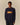 A person in the Rowing Blazers Cotton NY Hot Dog Sweater Navy, featuring “NEW YORK” on the front, stands against a plain beige background.