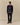 A man wears the Rowing Blazers x Babar Babar in New York Cotton Sweater Navy over a pink shirt, with dark jeans and brown shoes, standing against a plain beige background.