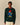 A man wears a Rowing Blazers x Paddington™ Paddington Cashmere Sweater Green, a dark green cashmere blend crewneck sweater featuring an intarsia knit of Paddington Bear in his blue coat and red hat, holding a jar of marmalade.