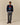 A person wearing a navy blue Rowing Blazers x Paddington™ Paddington Bear Cashmere Sweater over a button-down shirt, grey trousers, and brown loafers.