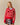 A person wearing the Warm & Wonderful Diana Edition Cotton Linen V-Neck Sheep Sweater Red over a blue collared shirt and plaid skirt, standing against a plain beige background.