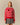 A person wears a Warm & Wonderful Wool Diana Edition Sheep Sweater Diana Red over a white collared shirt and a houndstooth pleated skirt, featuring a pattern of white sheep and a single black sheep on the chest.